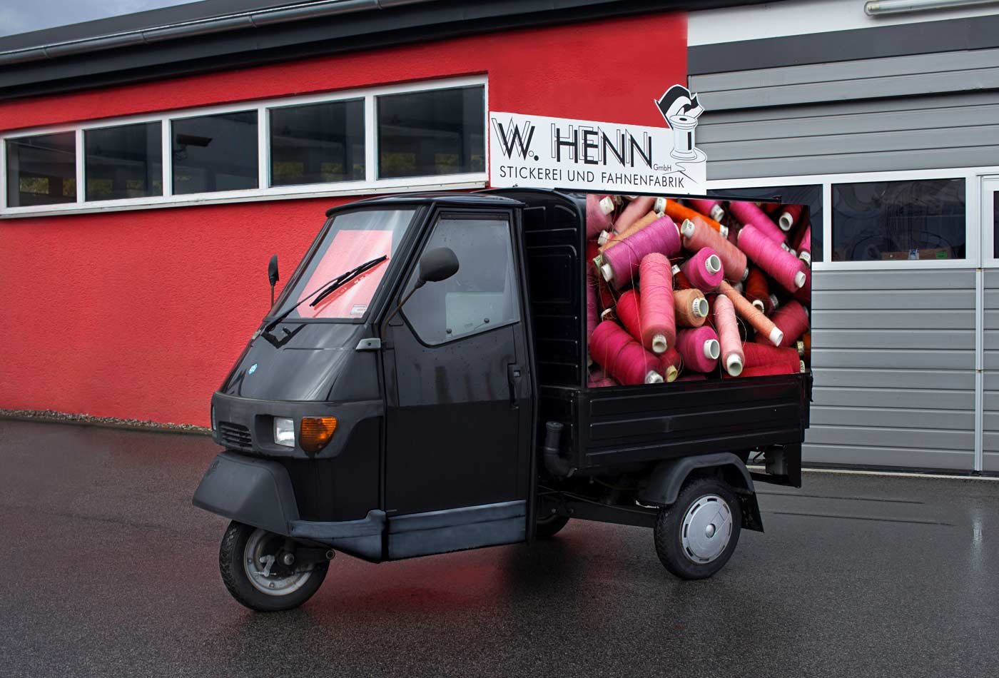 Dreirädiges Motorrad von Vespa, Ape genannt, überdacht, schwarz, hinten mit einem Koffer geschlossen. Seitlich ist ein Bild von bunten, in rot gehaltenen Garnspulen sichtbar, oben drauf ist ein weißes Schild mit dem Logo von Fahnen Henn und einer in Kontur geschnittenen Garnrolle